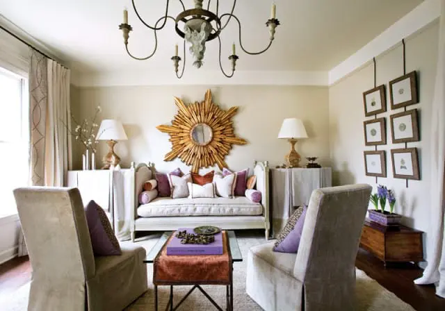 Living room with a cream-colored sofa, plush purple and rust pillows, a sunburst mirror, two upholstered chairs, and a dark wood chest with a planter.