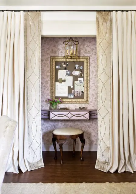 A vanity nook with floral wallpaper, a gold-framed bulletin board, a small upholstered stool, and cream curtains with decorative trim.