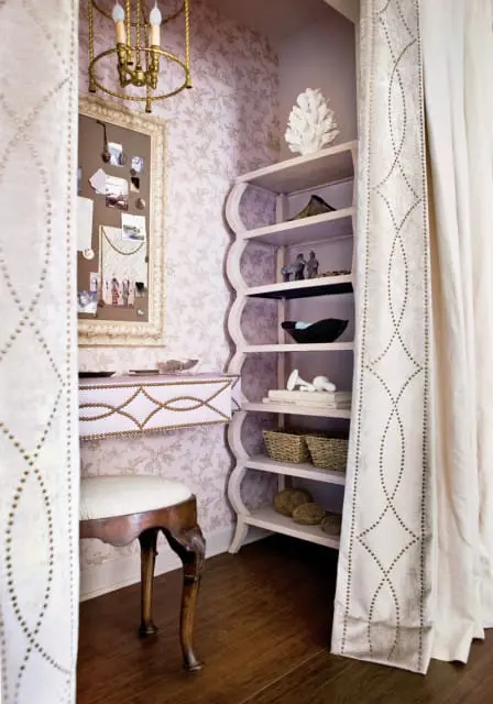 Vanity area with floral wallpaper, a corkboard, a pink vanity with gold trim, a wooden stool, and a scalloped shelf unit with decorative items.