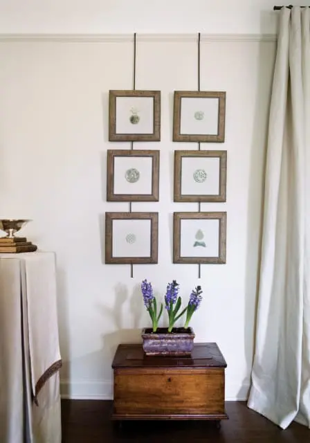 Wall with six framed botanical prints and a wooden chest with purple hyacinths. A draped table with books and a silver bowl is on the left.