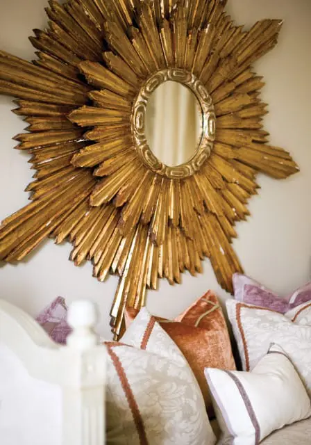 Gold sunburst mirror with an oval center above a collection of decorative pillows in shades of cream, rust, and lavender.