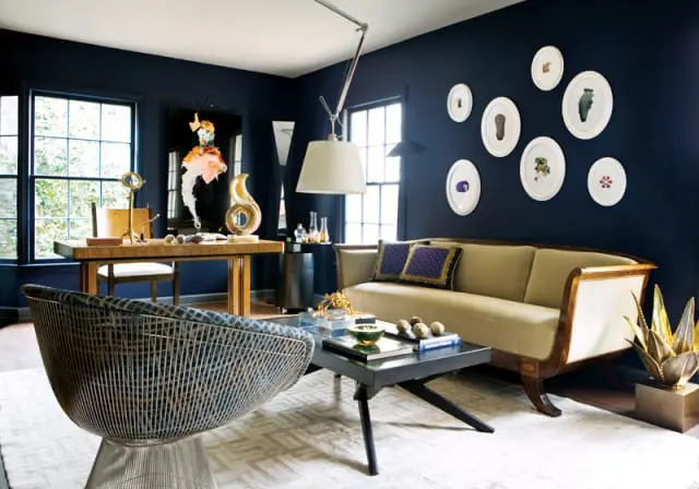 Dark blue living room with a light beige sofa, a patterned armchair, a wooden desk, and a black coffee table. Artwork and framed botanical prints adorn the walls.