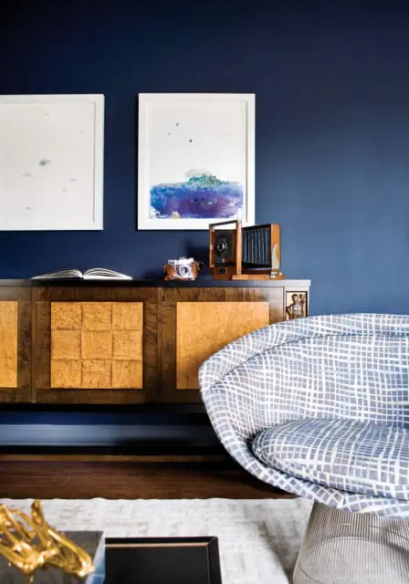 Dark blue walls with two framed abstract art pieces above a burl wood sideboard. A vintage camera sits on the sideboard next to an open book. A patterned armchair is in the foreground.