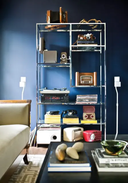 Glass and chrome shelving unit against a navy blue wall displaying vintage electronics including cameras, a record player, radios, and a rotary phone.