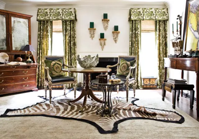 Living room with green floral curtains, antique wooden desk, two chairs with ornate pillows, and a zebra print rug over a cream rug.