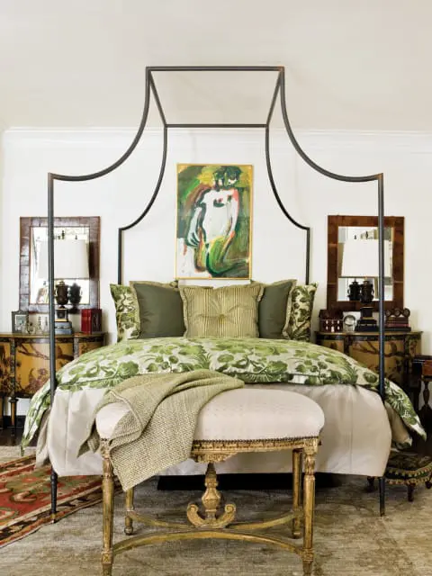Bedroom with a black canopy bed, green floral bedding, and an abstract painting above the headboard. A gold bench sits at the foot of the bed.