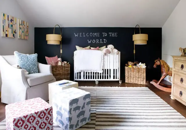 Nursery with a white crib, a white slipcovered armchair, and a black chalkboard wall with 'WELCOME TO THE WORLD' written in chalk.