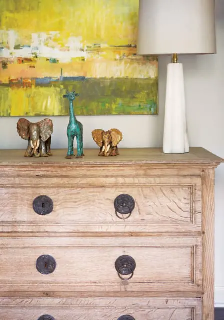 Light wood dresser with three drawers featuring ornate metal pulls, topped with two elephant figurines, a teal giraffe figurine, and a tall white lamp, against a backdrop of abstract yellow and green art.