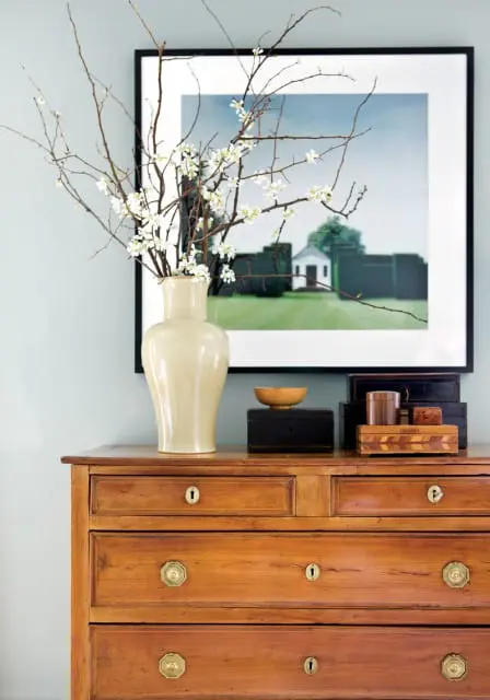 Light blue walls with a wooden dresser topped by a cream vase holding branches with white blossoms and decorative boxes.