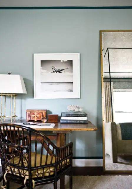 Desk with a bamboo chair, a framed black and white photograph of an airplane taking off, and a stack of books. A large floor mirror with a gold frame stands to the right.