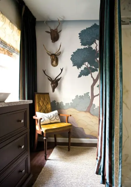 Bathroom with taxidermied deer heads mounted on a wall with a scenic mural, a velvet armchair, and dark wood vanity.
