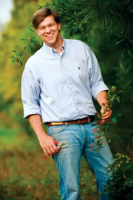 Man in light blue button-down shirt and jeans holding pruning shears and a branch with berries, smiling outdoors.
