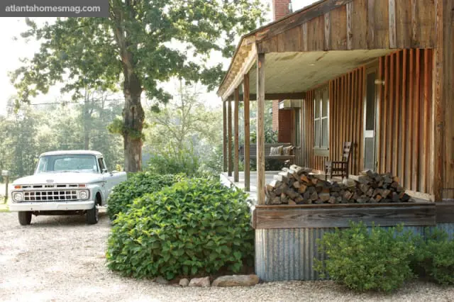 Rustic wooden house with a covered porch, stacked firewood, and a vintage white pickup truck parked on a gravel driveway.