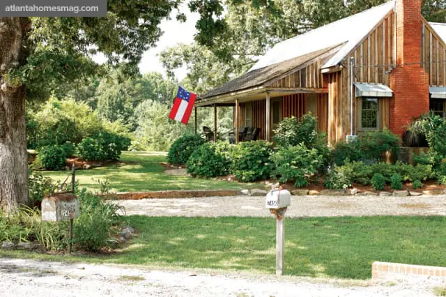 Rustic wooden house with a covered porch, brick chimney, and a flag flying. Surrounded by lush green trees and manicured bushes.