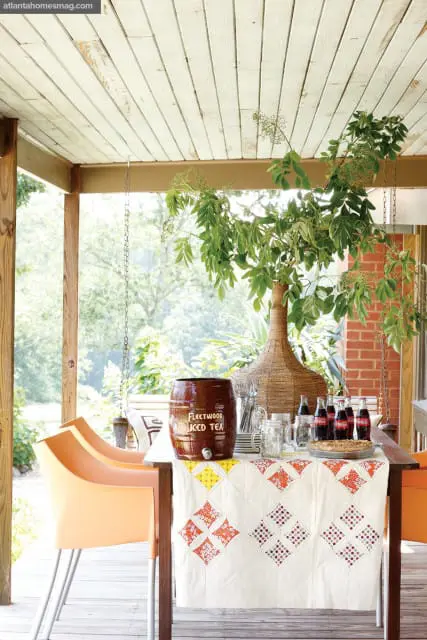 Covered porch with white wood-paneled ceiling, wooden posts, and a table set for a gathering with a large brown iced tea dispenser, bottles of Coca-Cola, mason jars, and a pie.