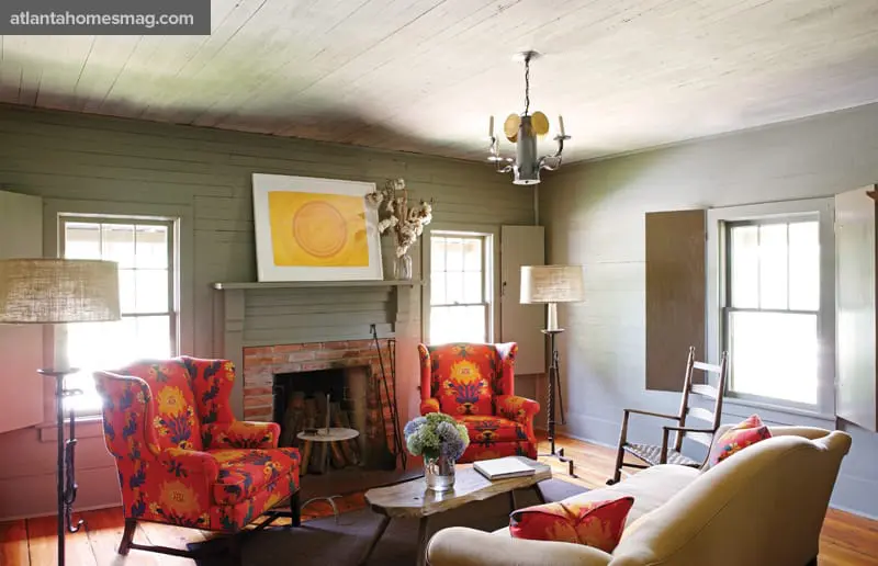Living room with sage green paneled walls, a brick fireplace, two wingback chairs with red and blue floral upholstery, and a light-colored sofa.