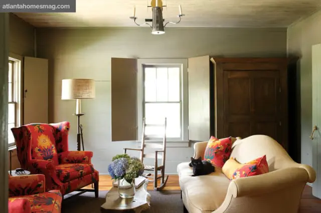 Living room with light gray paneled walls, a cream sofa with floral pillows, a red wingback chair with floral upholstery, a wooden armoire, and a floor lamp.