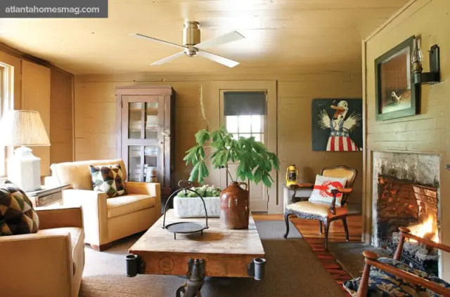 Living room with tan walls and ceiling, featuring two tan armchairs, a rustic wooden coffee table with metal accents, a vintage glass-front cabinet, and a stone fireplace with a fire burning.