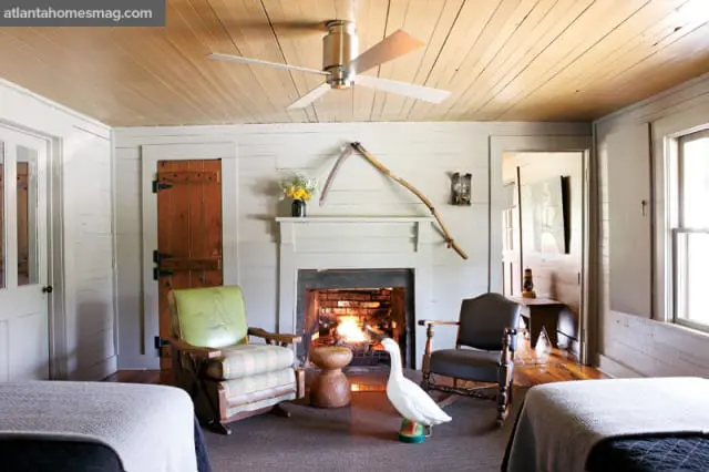 Bedroom with wood-paneled ceiling, fireplace with a scythe above, two rocking chairs, and a white goose statue.