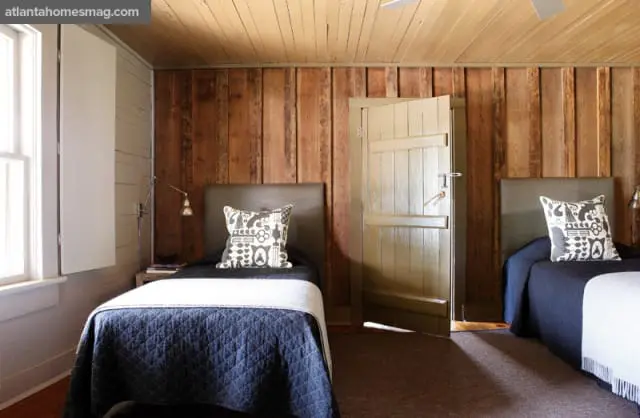 Bedroom with wood-paneled walls and ceiling, two beds with dark blue quilts and patterned pillows, and a light green door slightly ajar.