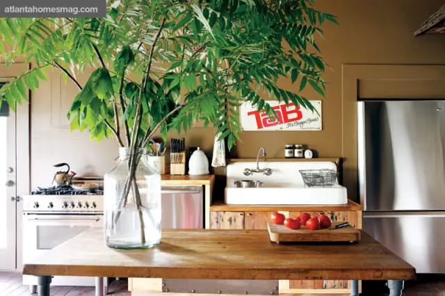 Brown kitchen with stainless steel appliances, a large farmhouse sink, and a rustic wooden island topped with a vase of greenery and a cutting board with tomatoes.