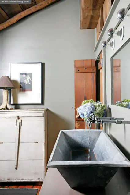 Rustic bathroom with a galvanized metal trough sink, vintage cabinet, wooden barn door shutter, and framed artwork on a pale green wall.