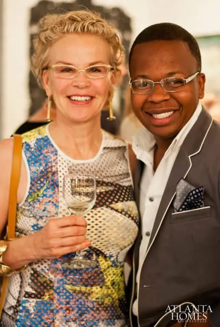 Woman with curly blonde hair wearing glasses and a colorful patterned dress holding a wine glass, next to a man wearing glasses and a grey suit jacket with a pocket square.