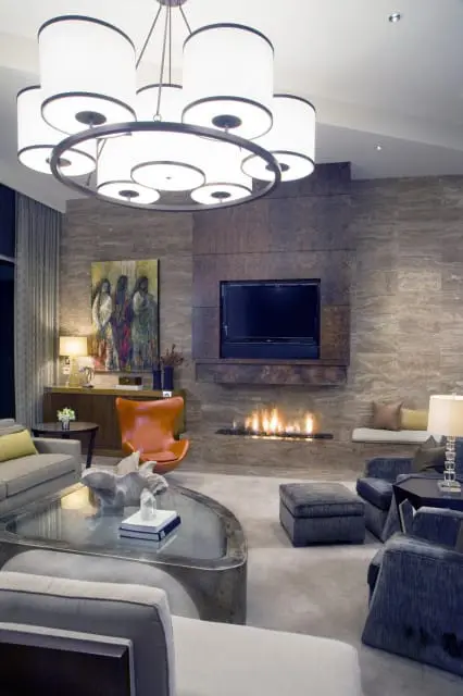 Modern living room with a large circular chandelier, a fireplace with a television above, a tan armchair, and gray sofas.