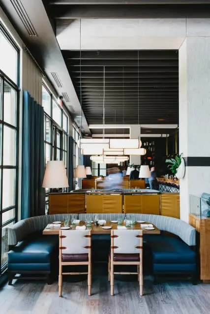 Restaurant dining area with curved blue banquette seating, wooden tables set for dining, and yellow upholstered booth backs. Large windows with blue drapes line the left wall.