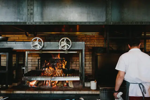 Commercial kitchen with a large brick wood-fired grill, metal hood, and a chef in a white uniform standing with his back to the camera.