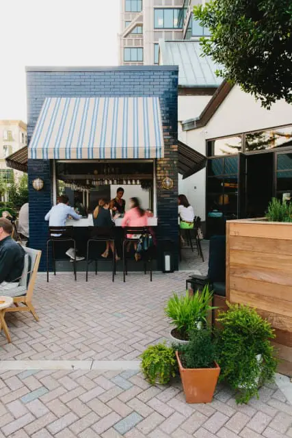 Outdoor bar with blue brick facade and striped awning, patrons seated at the counter, and potted plants on a brick patio.