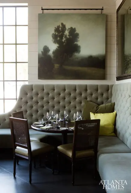 Dining area with a tufted grey banquette, dark wood table set for dinner, cane-backed chairs, and a large landscape painting above.