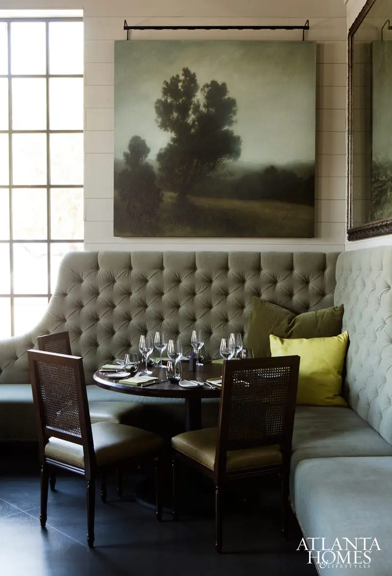 Dining area with a tufted grey banquette, dark wood table set for dinner, cane-backed chairs, and a large landscape painting above.