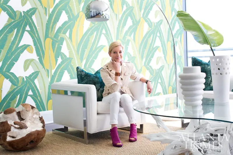 Woman in white armchair against banana leaf wallpaper, wearing a tan jacket, white pants, and magenta heels. A glass coffee table with white sculptural bases sits in front.