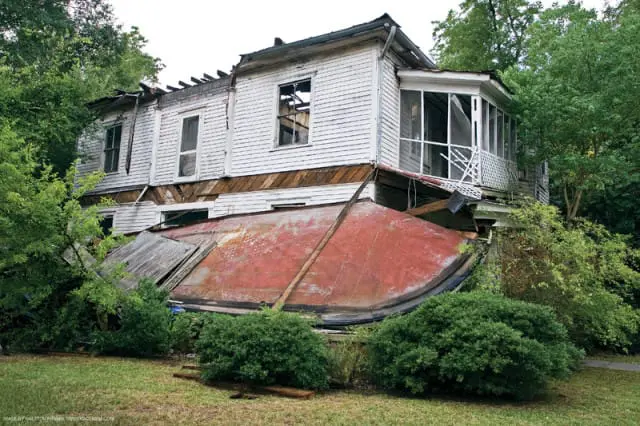 Dilapidated white clapboard house with a collapsed section revealing a red metal roof underneath, surrounded by overgrown greenery.