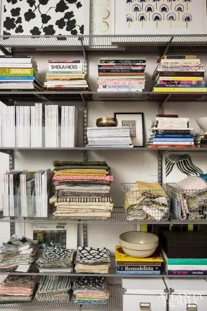 Metal shelving unit filled with stacks of books, magazines, folded fabrics, and decorative items. Notable books include 'Sheila Hicks: Bead Embroidery', 'Marimekko: The Drawing Book', and 'Remodelista'.