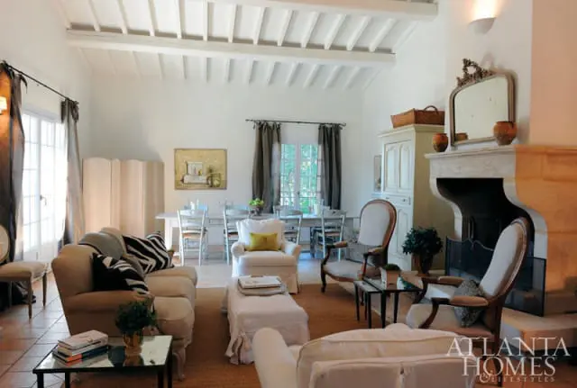 Living room with a stone fireplace, white beamed ceiling, neutral-toned furniture including sofas and armchairs, and a dining area visible in the background.