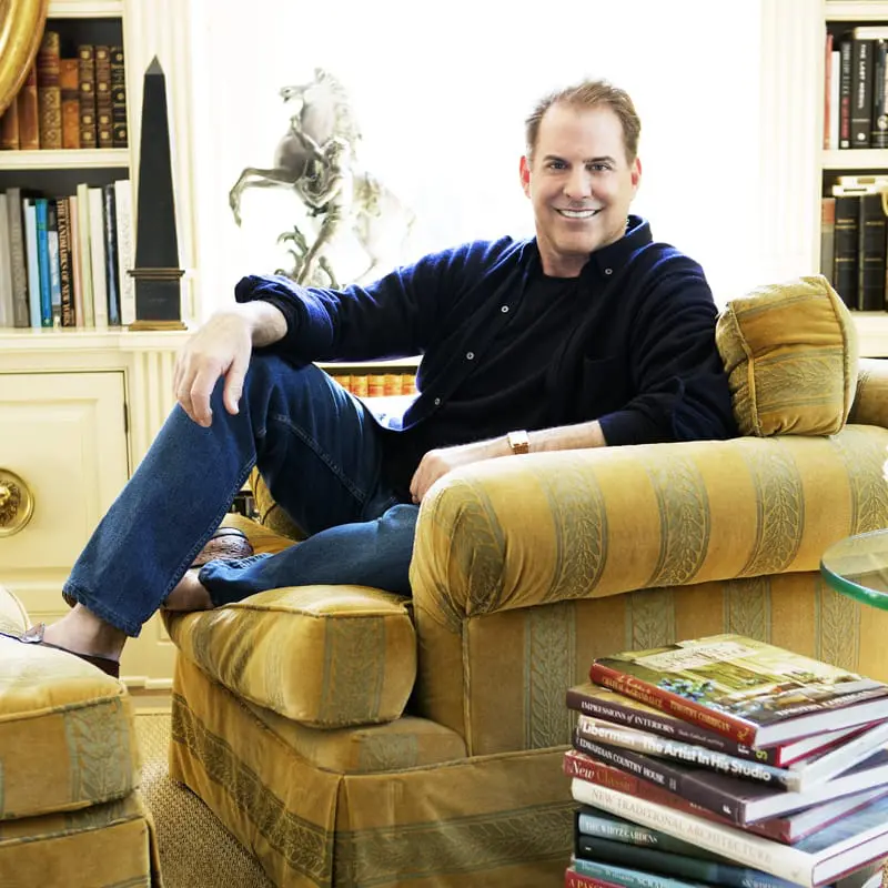 Man in dark blue sweater and jeans sitting in a gold upholstered armchair, with bookshelves and a sculpture in the background.