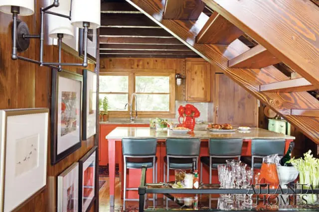 Kitchen with wood-paneled walls, a coral island with granite countertop, and dark green bar stools. A wooden staircase with exposed beams is overhead.