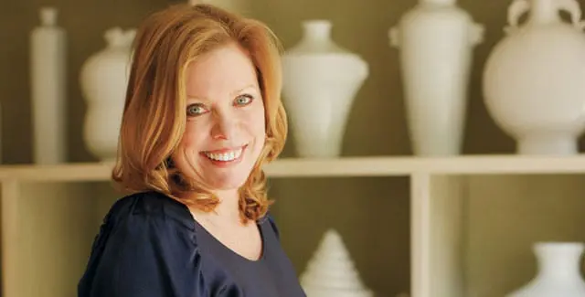Woman with auburn hair and blue eyes smiling, wearing a navy blue top, with shelves of white vases in the background.