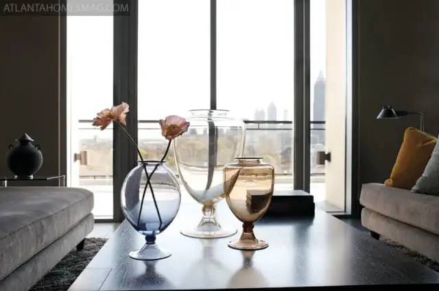 Living room with a dark wood coffee table featuring three decorative glass vases and two flowers, overlooking a city skyline through a large window.