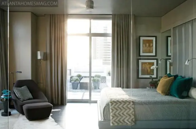 Modern bedroom with gray walls, a large window overlooking a balcony, a bed with teal and gold pillows, and a dark gray armchair with an ottoman.