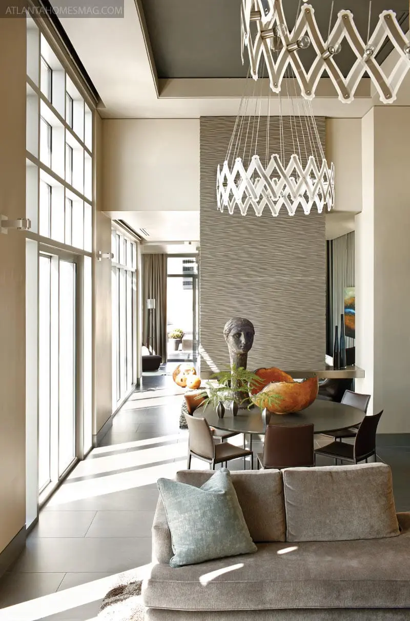 Modern dining room with textured gray wall, round table with chairs, and a large sculptural chandelier. A sofa with a blue pillow is in the foreground.
