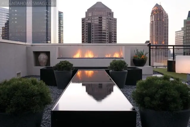 Rooftop patio with a modern gas fireplace, reflective black table, potted evergreen shrubs, and city skyline view at dusk.