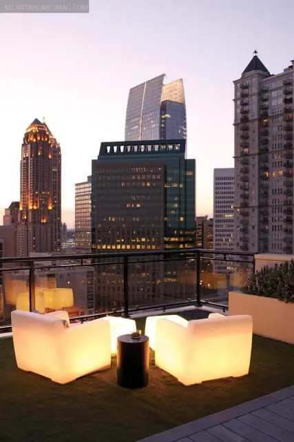 Rooftop patio with glowing white chairs and a dark cylinder table, overlooking a city skyline at dusk.