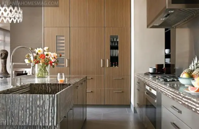 Modern kitchen with wood-paneled cabinets, a granite waterfall island with a farmhouse sink, and stainless steel appliances. A vase of flowers sits on the island.