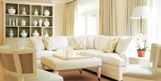 Living room with a sectional sofa in cream upholstery, patterned throw pillows, and a tufted ottoman. A built-in bookshelf filled with white ceramic vases is visible.
