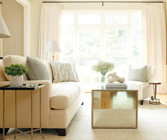 Cream sofa and armchair in a living room with large windows, cream drapes, a mirrored coffee table, and a green floral arrangement.