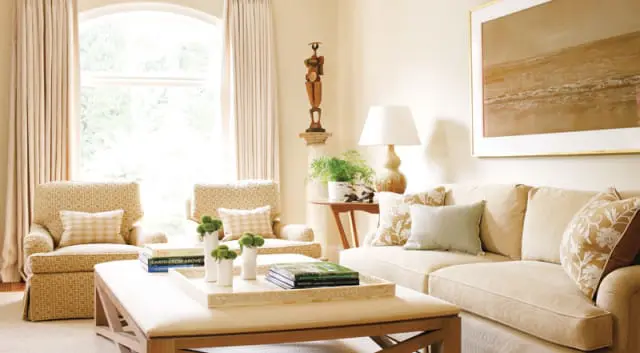 Living room with two patterned armchairs, a light beige sofa, and a large upholstered ottoman coffee table. Neutral tones dominate, with accents of green in plants and pillows.