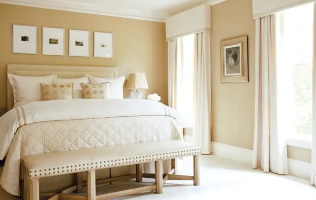 Bedroom with a king-sized bed dressed in white linens, a quilted duvet, and decorative pillows. A tufted bench sits at the foot of the bed. Walls are a warm beige, adorned with framed artwork.